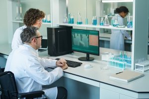 From Discovery to Translation Side view of two professional virologists in lab coats discussing virus cell on computer screen while one of them sitting in wheelchair